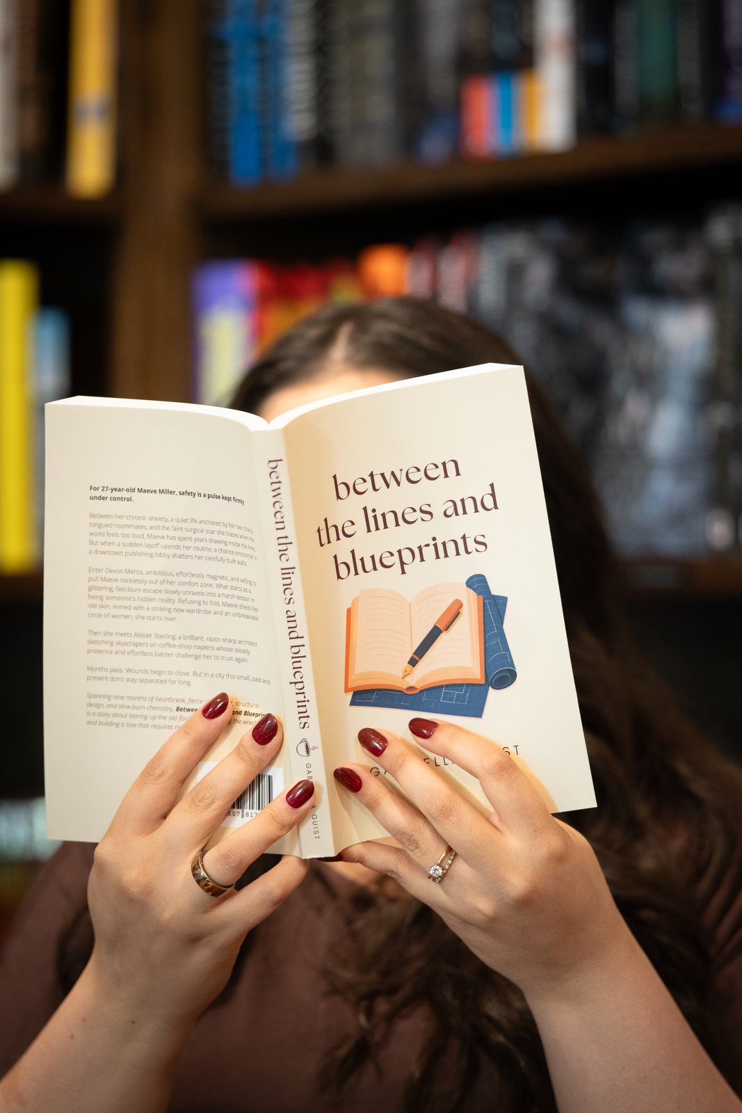 Gabriella holding Between the Lines and Blueprints open in front of her face, bookshelves behind